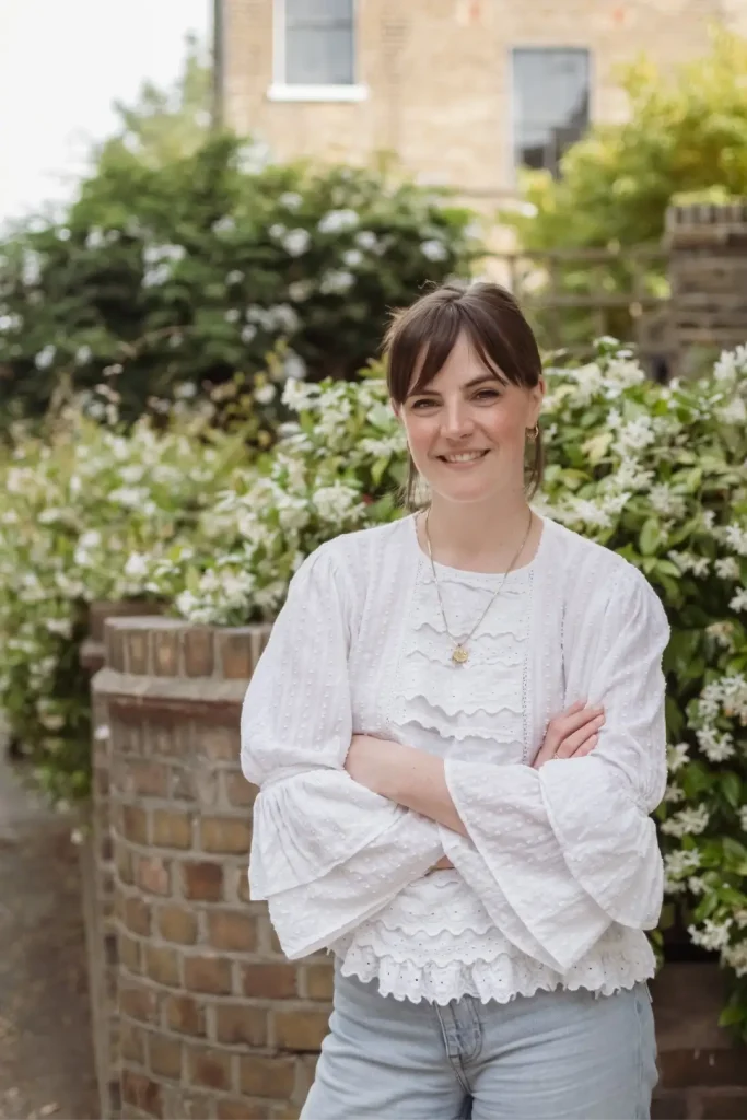 A woman with brown hair and a fringe stands with her arms crossed, smiling at the camera. She wears a white broderie anglaise blouse with bell sleeves and a delicate gold pendant necklace, standing in front of a low flowery brick wall.