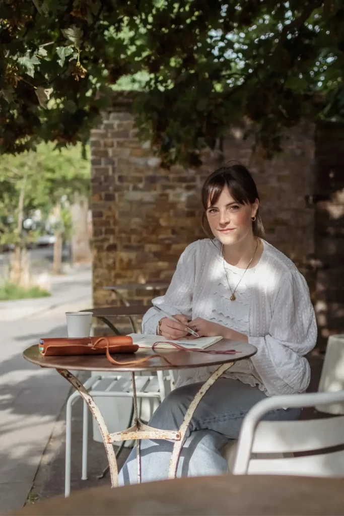 A woman with brown hair and a fringe sits at a small round café table outdoors, looking up from an open leather-bound notebook with a pen in hand. She wears a white broderie anglaise blouse and jeans, with dappled sunlight filtering through the tree canopy above and a takeaway coffee cup beside her on the table.