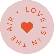 A circular pink sticker with red writing, spelling the words "Love is in the air." There is an icon of a heart in the centre of the sticker.