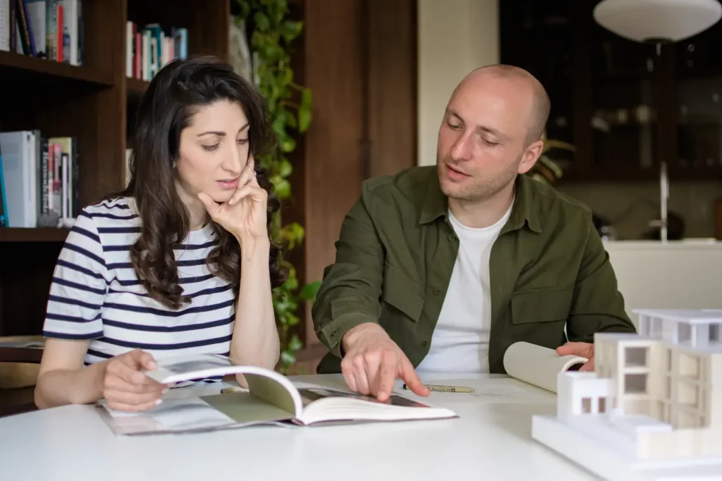 Two architects sit at a white table reviewing an open book together, with a man in an olive shirt pointing at a page while a woman in a striped top looks on. White architectural models and a dark wooden bookcase fill the background.