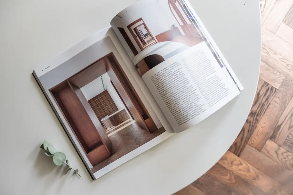 An open architecture magazine lies on a white round table beside a sprig of eucalyptus, its pages showing interior photographs and Romanian text, with parquet flooring visible alongside.