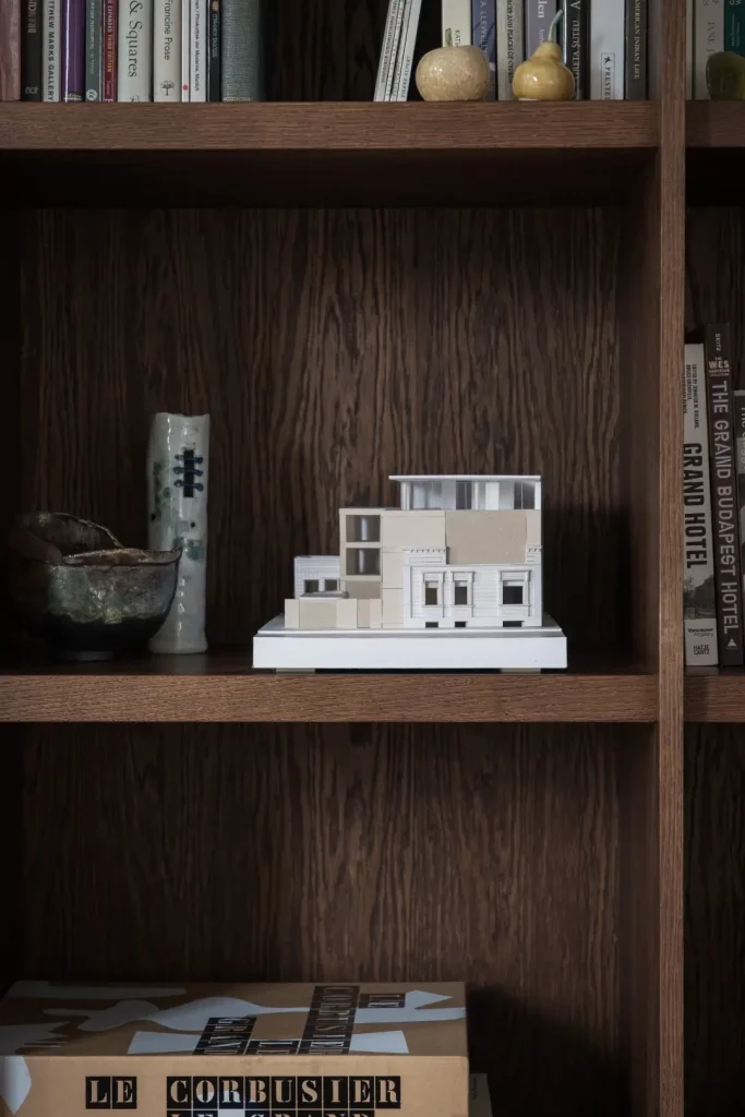 A detailed white architectural model of a house sits on a shelf within a dark wood bookcase, surrounded by ceramic pieces and books including titles on Le Corbusier and the Grand Budapest Hotel.