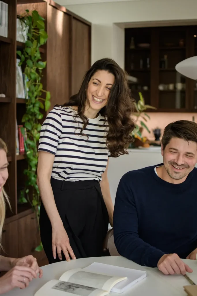 Three colleagues share a light-hearted moment around a table, with a woman in a striped top standing and laughing between a seated blonde woman and a man in a navy jumper. Trailing houseplants and wooden cabinetry frame the scene.