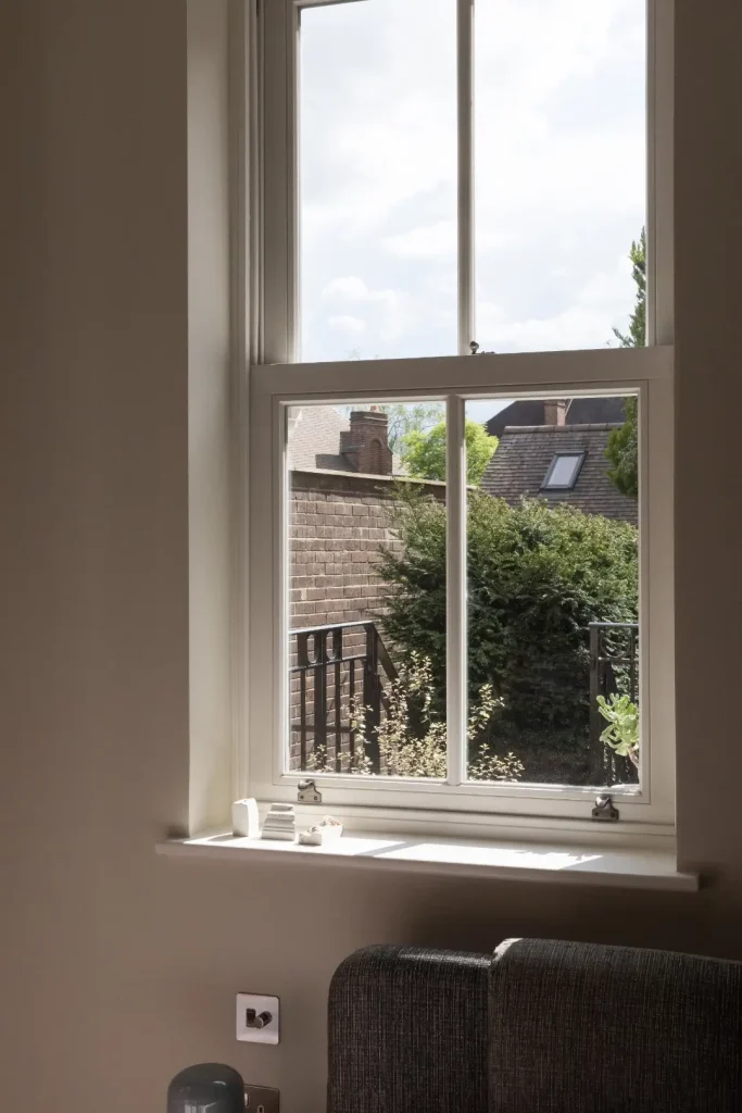 A white-framed sash window set in a warm beige wall, looking out onto a brick garden wall, tiled rooftops and greenery, with small ceramic objects on the sill and sunlight falling across it.