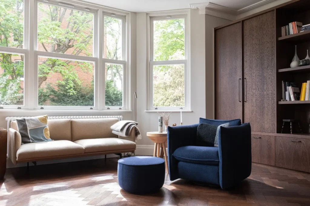 A bright living room with a beige sofa beneath tall windows overlooking trees, a navy armchair and matching round pouffe, and built-in dark wood cabinetry with bookshelves. Herringbone parquet flooring runs throughout.