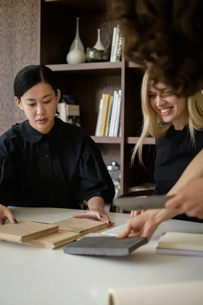 Two architects sit at a table examining timber and stone material samples, one in a black blouse looking down attentively and the other smiling, with a wooden bookcase behind them.