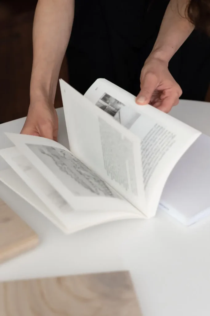 A person in black holds open an architecture book above a white table, fanning through its pages of text and black-and-white images.