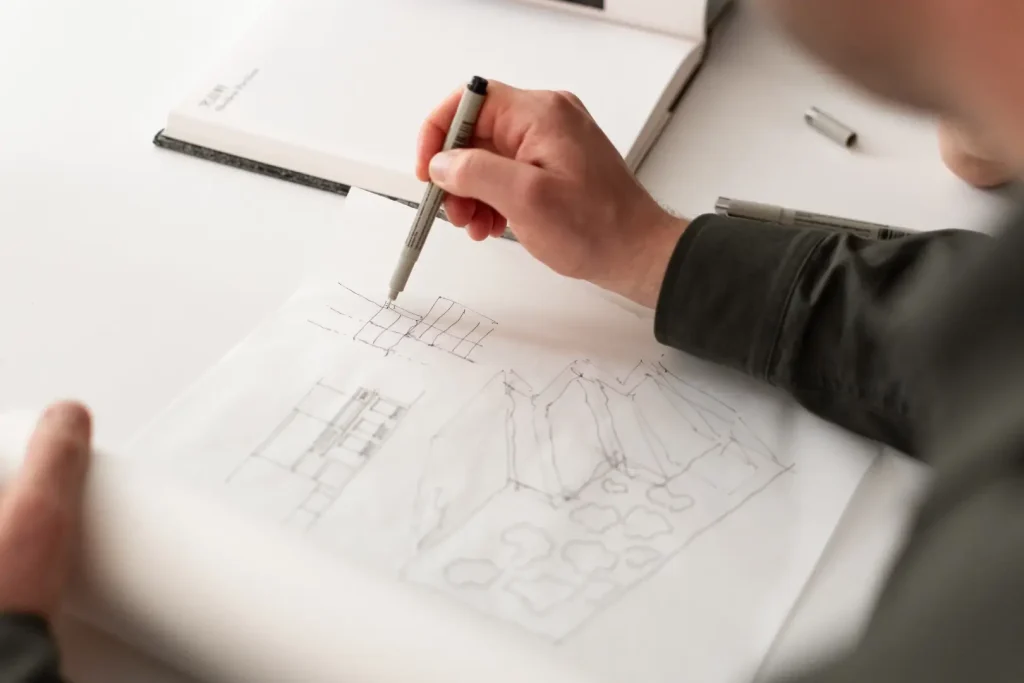 A close-up of a hand sketching building elevations and a perspective drawing in fine black pen on white paper, with a notebook and pens nearby.
