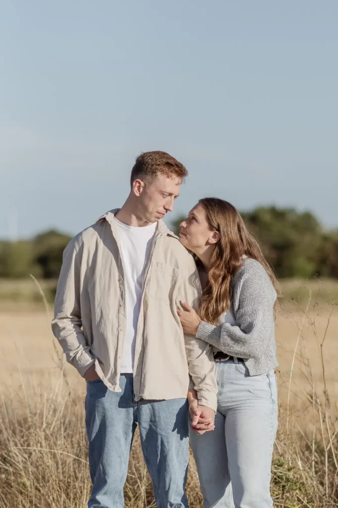 An engaged couple stand close together in a sunlit field, holding hands as she looks up at him and he glances down, both bathed in warm afternoon light.