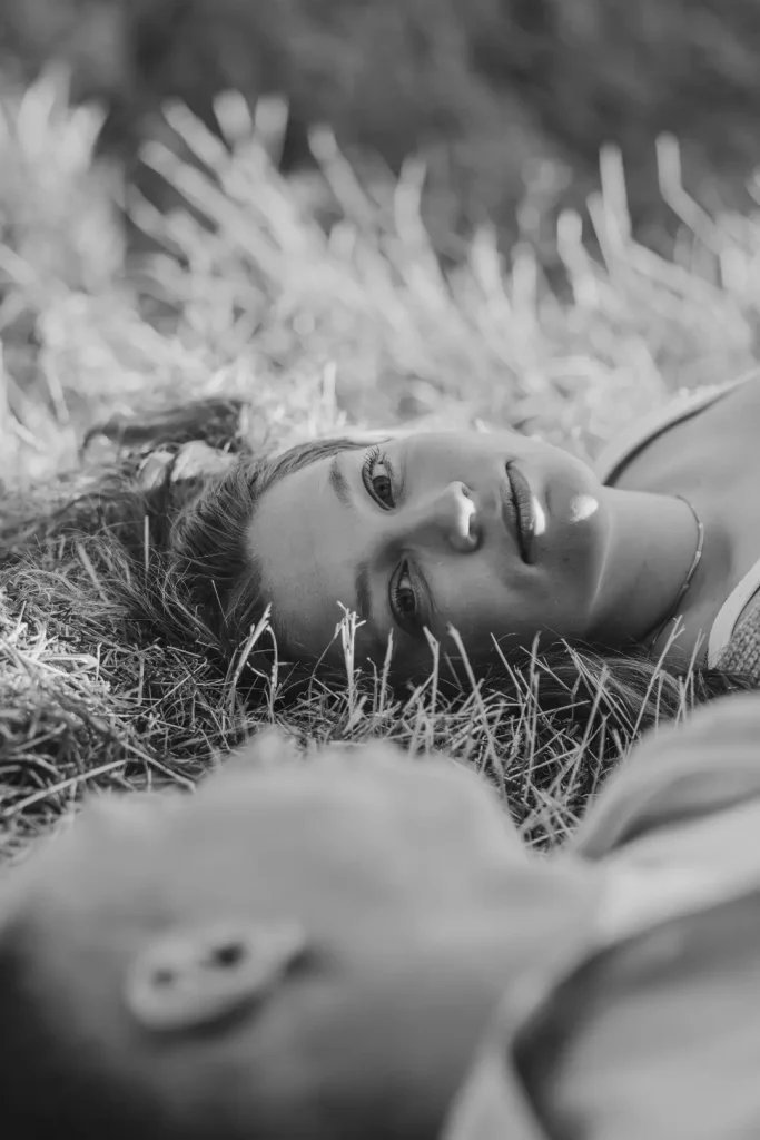 A soft black-and-white portrait of a woman lying back in long grass, smiling gently up at the camera, with her partner blurred in the foreground.