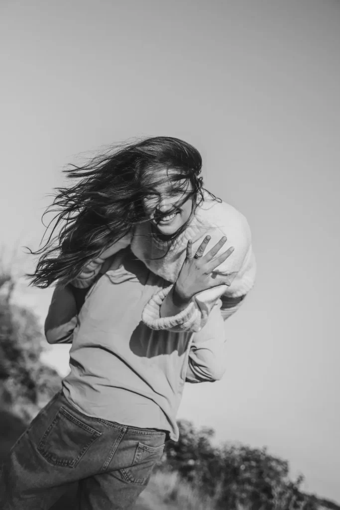 Black-and-white photo of a woman laughing as her hair blows across her face in the wind, as she is carried by her partner. Her engagement ring visible on her finger.