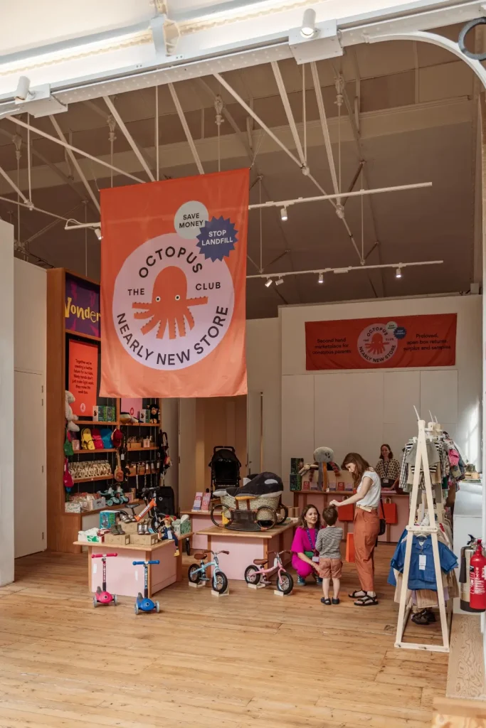 Wide view of The Octopus Club Nearly New Store at the Young V&A, with a large orange "Save Money, Stop Landfill" banner suspended from the high pitched ceiling. Pink display tables hold scooters, balance bikes and pre loved toys, while a clothing rail and shelves of accessories line the walls.