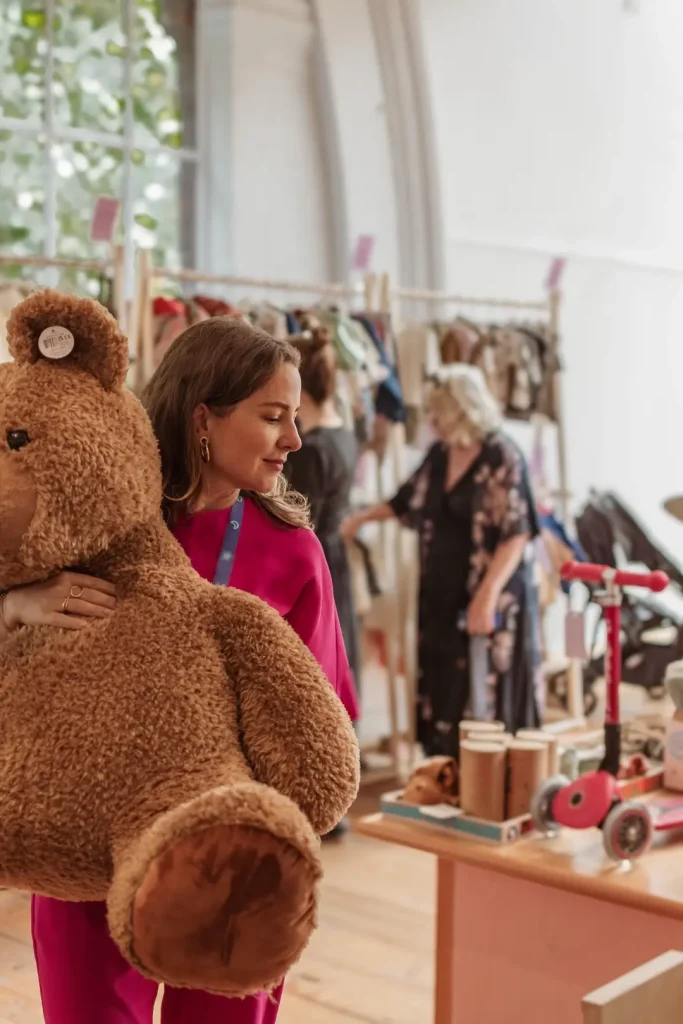 A woman in a pink top carries a large brown teddy bear across the store, with other shoppers browsing clothing rails and a red scooter on a display table behind her.