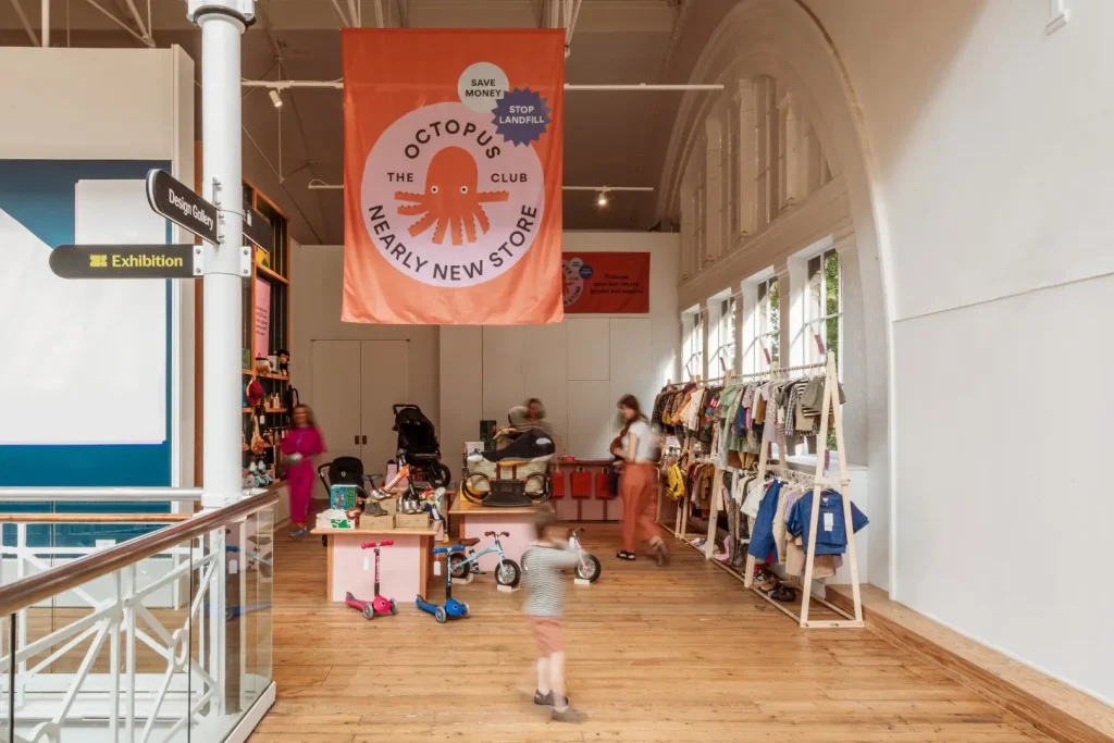 Wide interior shot of the Nearly New Store entrance with the large orange Octopus Club banner overhead, families browsing rails of pre loved clothing on the right and a child running past in motion blur.