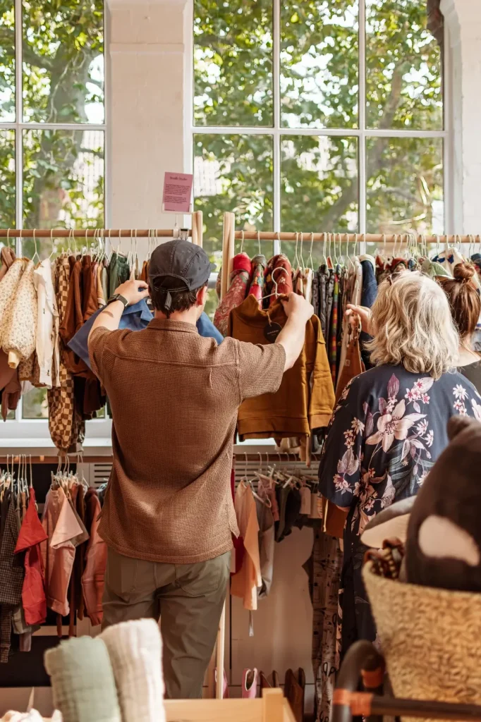 A man in a brown waffle shirt and cap browses a wooden rail of pre loved children's clothing alongside two other shoppers, with sunlit windows and trees behind.