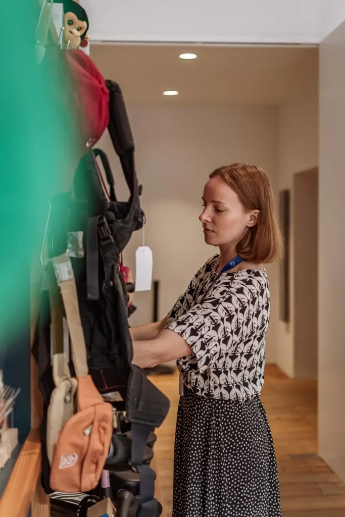 A staff member with a bob haircut, wearing a black and white patterned blouse and polka dot skirt, sorts through pre loved baby carriers and accessories displayed on a wall.