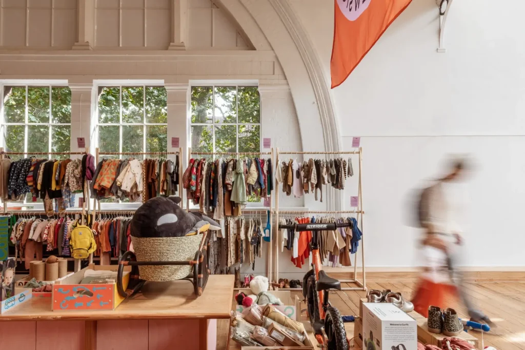 Interior view of a pre loved store, with rails of children's clothing lined up beneath an arched ceiling and tall windows, a large plush orca in a Moses basket on a wooden display table, and a shopper walking past in motion blur.