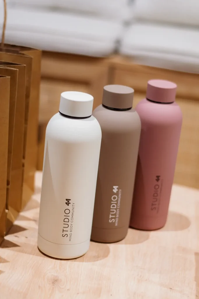 Three insulated Studio 44 water bottles in cream, taupe and dusky pink, lined up on a wooden surface beside kraft paper bags. Each bottle is branded with "Studio 44 - Mind Body Community."